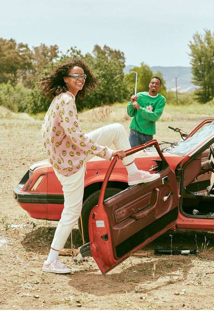 A woman in a patterned top and white pants sits on the door of a damaged orange car in an open, grassy field with another man nearby.
