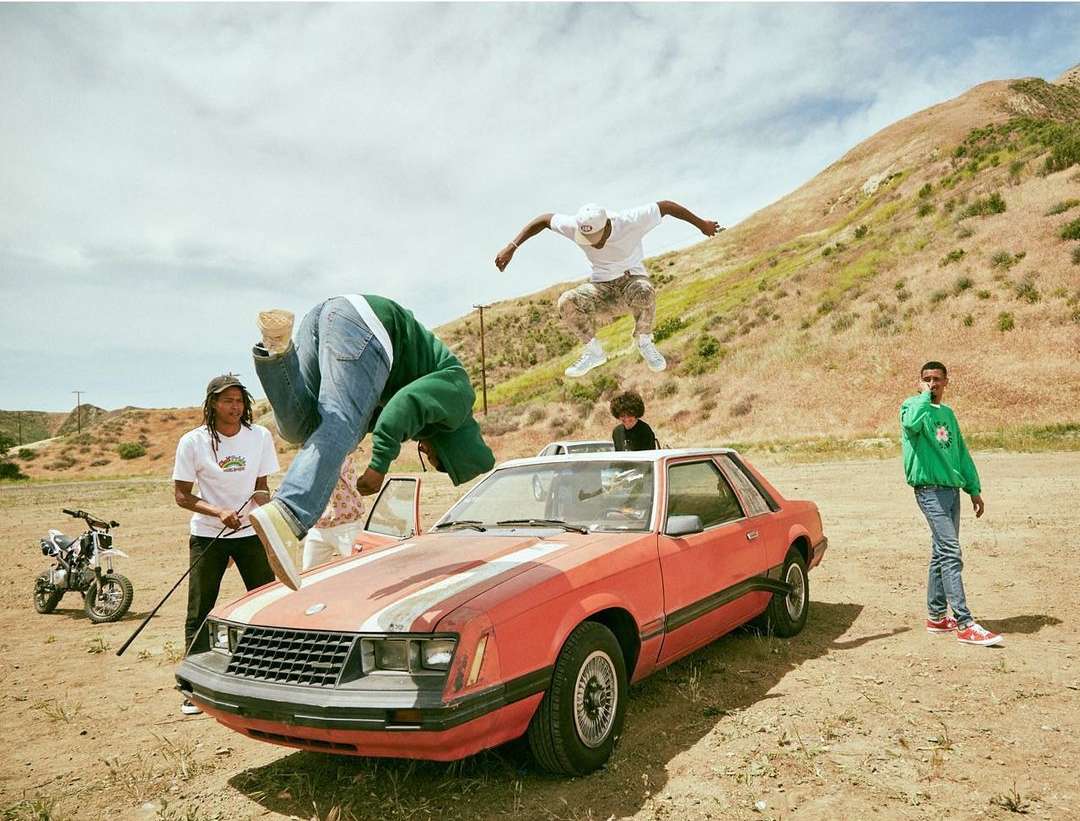 A group of four young adults near a red car, two are jumping in the air in a desert landscape with a dirt bike visible to the left.
