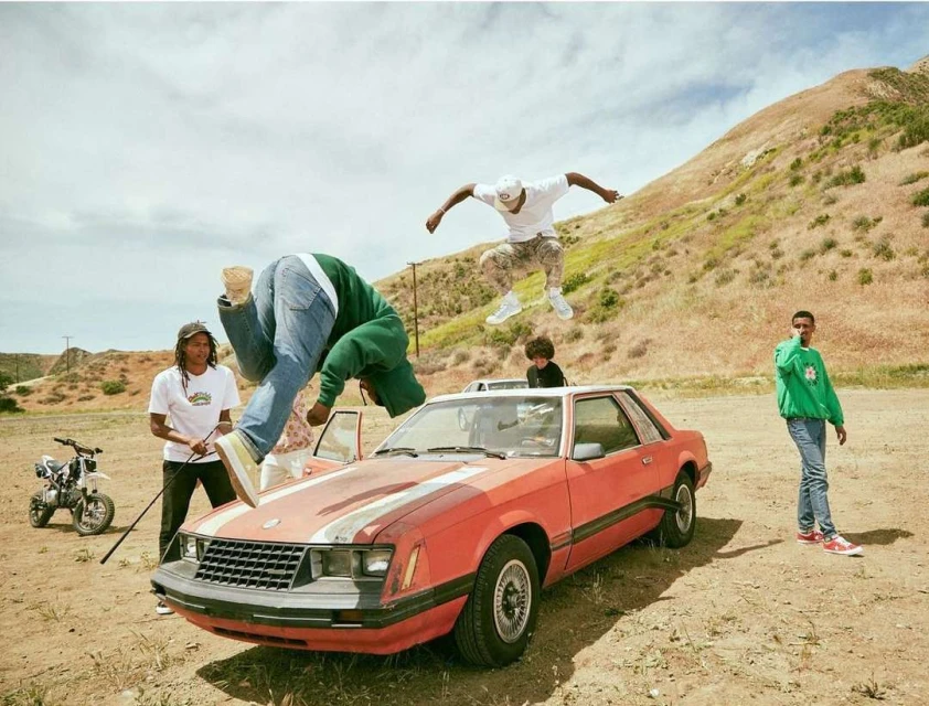 A group of four young adults near a red car, two are jumping in the air in a desert landscape with a dirt bike visible to the left.