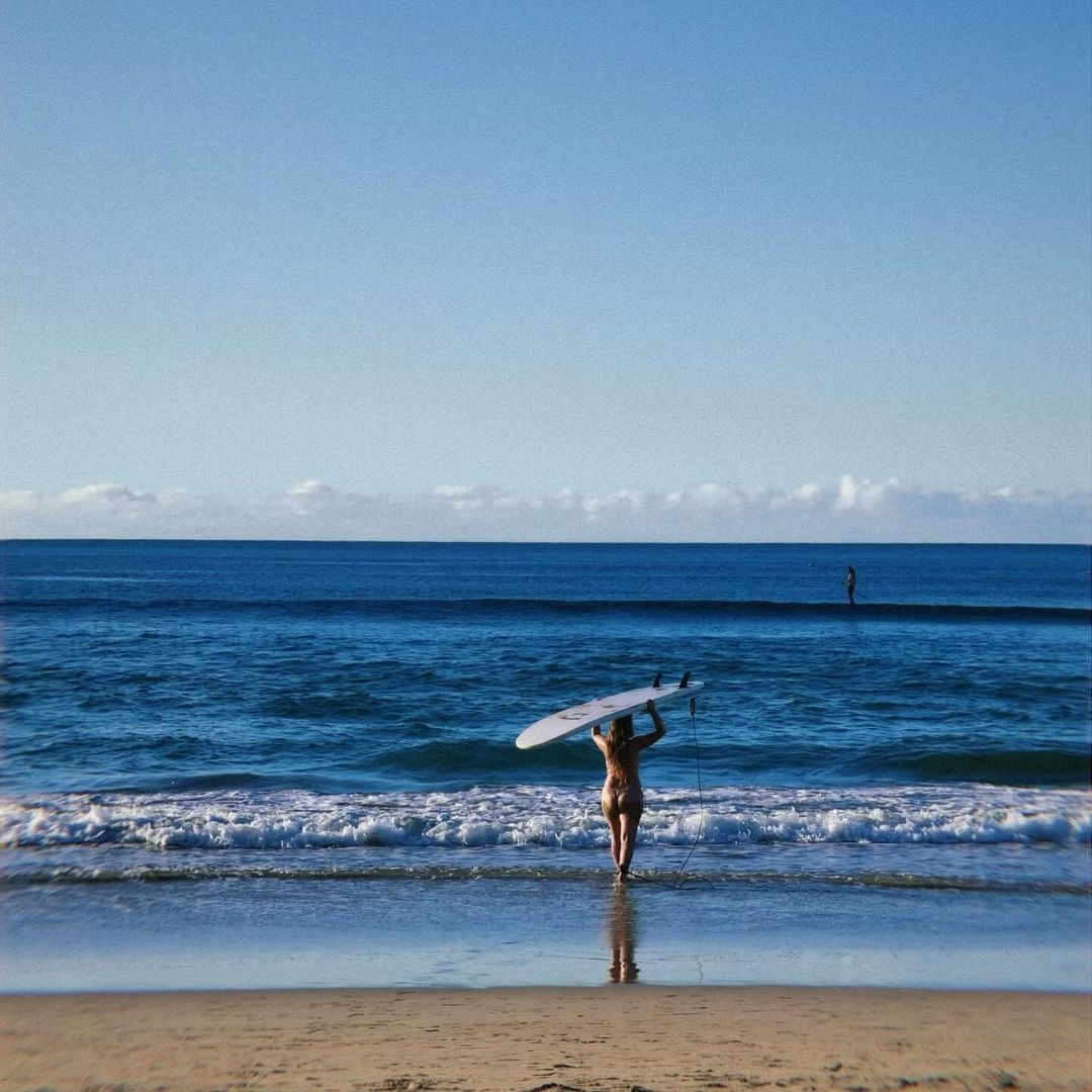 Woman with surfboard on her head walks into ocean at beach on a sunny day; another surfer visible in the background standing in the water.