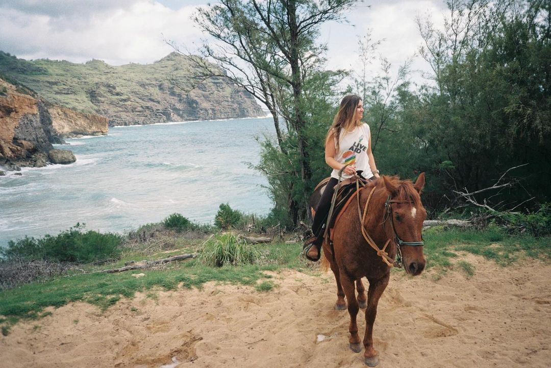 A woman rides a horse along a sandy path near the ocean, with cliffs and greenery in the background, on a bright day.