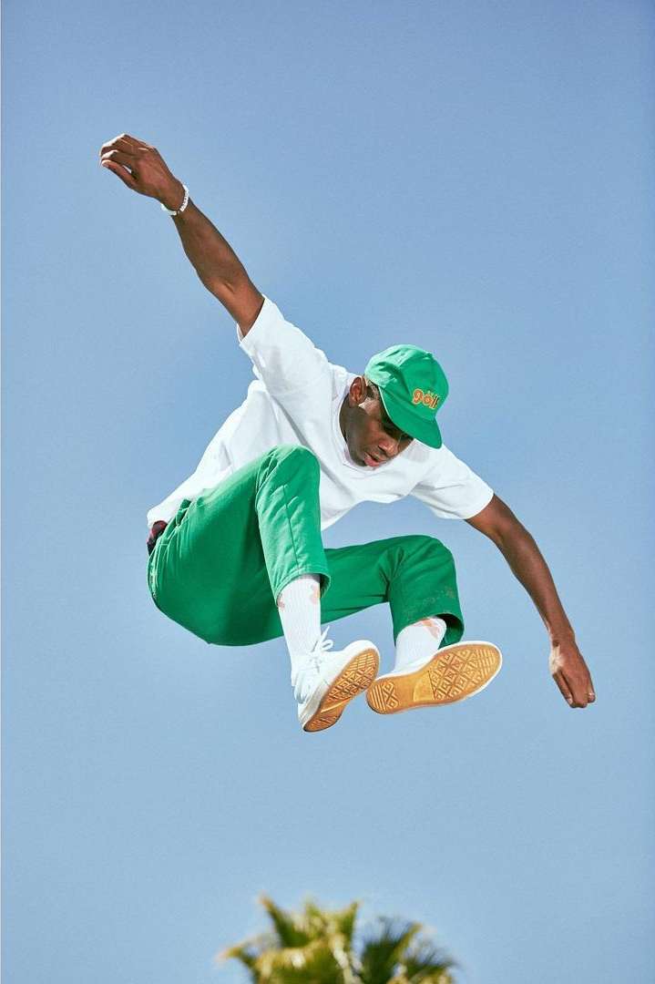 An African-American man in a white t-shirt, green pants, and a green cap jumps high in the air, showcasing casual fashion against a blue sky.