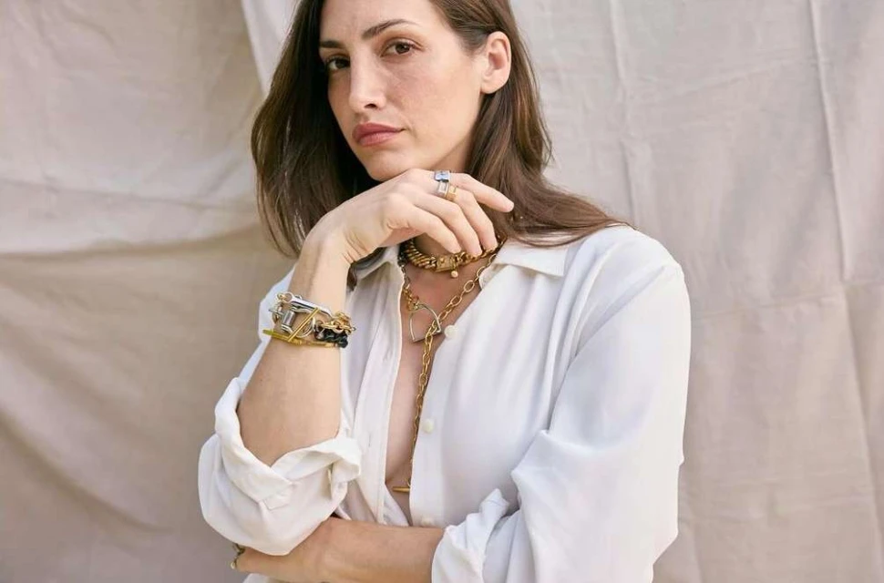 Portrait of a woman in a white shirt wearing multiple gold chain necklaces, gold rings, and a mixed-metal bracelet indoors.