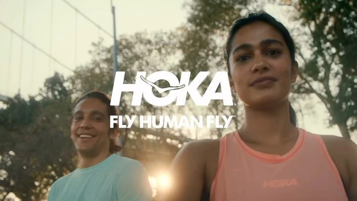 Outdoor shot of an adult woman and man smiling with the HOKA logo and slogan overlaid on the image, on a sunny day.