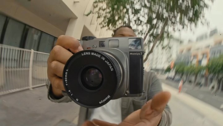 Man holding a Mamiya 7 medium format film camera in an urban setting with trees and buildings visible in the blurred background.