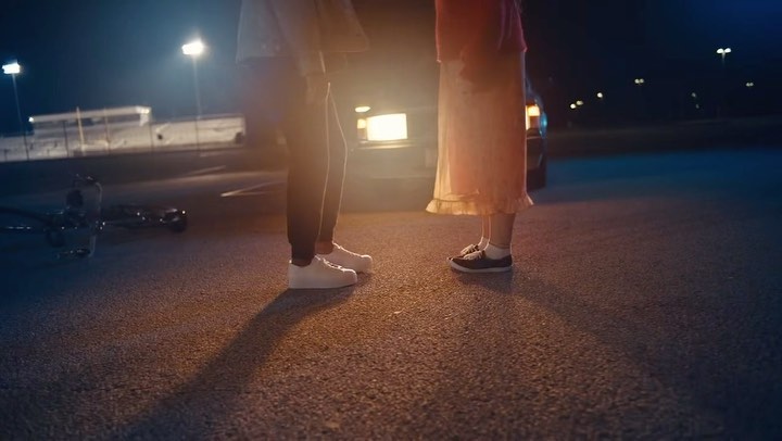 Low-angle shot of a couple standing on an empty asphalt lot at night, illuminated by car headlights and street lights.