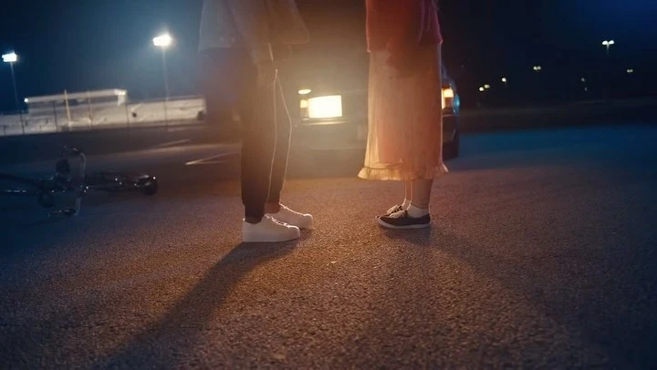 Low-angle shot of a couple standing on an empty asphalt lot at night, illuminated by car headlights and street lights.