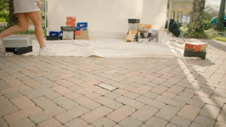 Woman walks past stacked shoeboxes in an outdoor setting with brick paving and leafy trees, creating a casual lifestyle image.