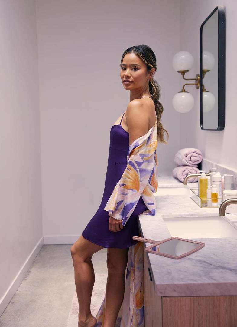 Woman in a purple slip dress poses in a bathroom with neutral colors and marble counters. She is wearing a floral print shawl.
