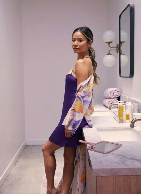 Woman in a purple slip dress poses in a bathroom with neutral colors and marble counters. She is wearing a floral print shawl.