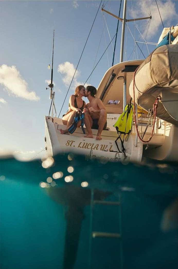 A couple kisses on the stern of a sailboat, with the boat partially submerged, capturing the waterline and the underwater view.