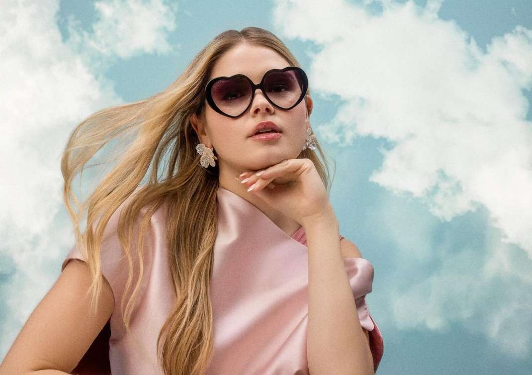 Blonde woman poses in a light pink dress with heart-shaped sunglasses against a cloudy sky. She is wearing flower earrings.