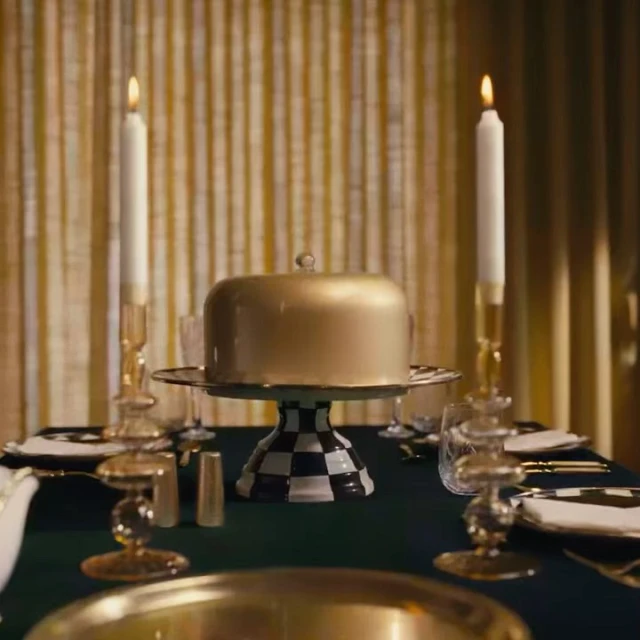 Elegant table setting featuring a gold covered cake on a black and white checkered stand, flanked by lit candles in gold holders.