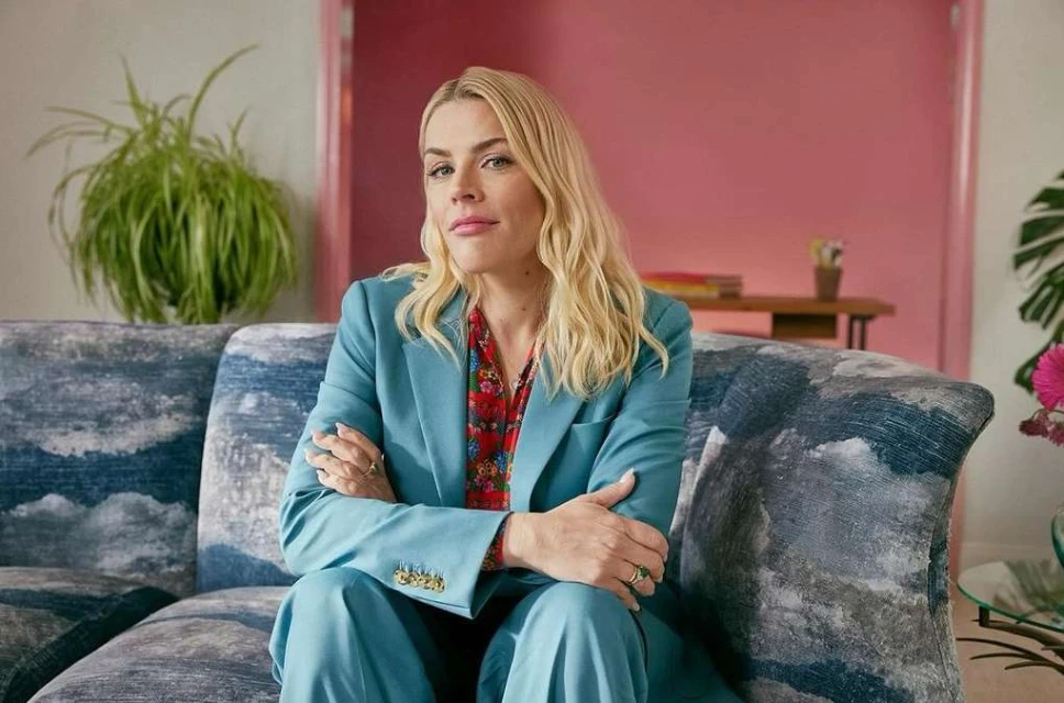 Blonde woman in a blue suit sitting on a patterned sofa indoors, with a pink door and houseplant visible in the background.
