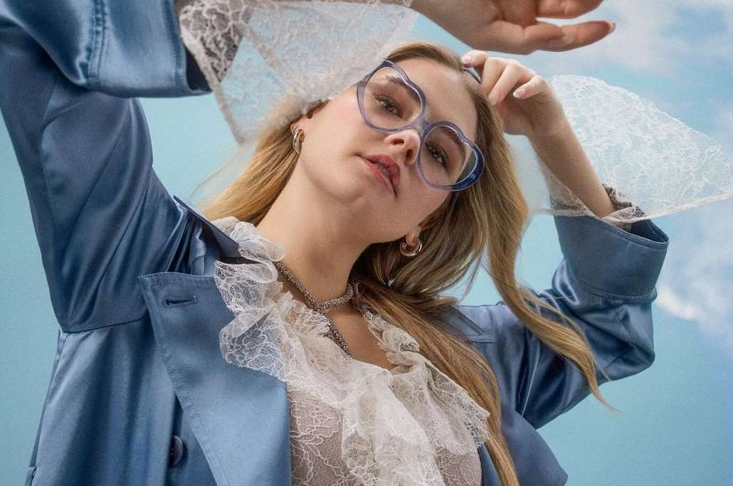 Blonde woman wearing glasses and a blue jacket poses against a light blue background, showcasing fashion and beauty in a close-up studio shot.