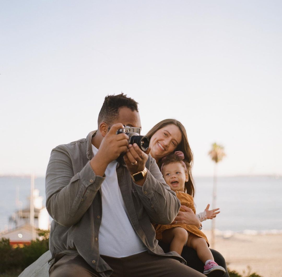 Family of three poses together near the ocean, father takes a photo with a film camera, mother and baby smile on a sunny day.