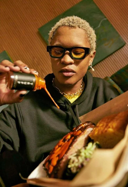 Person with short blonde hair in black glasses pouring orange sauce on ribs with coleslaw inside a brown paper food container.