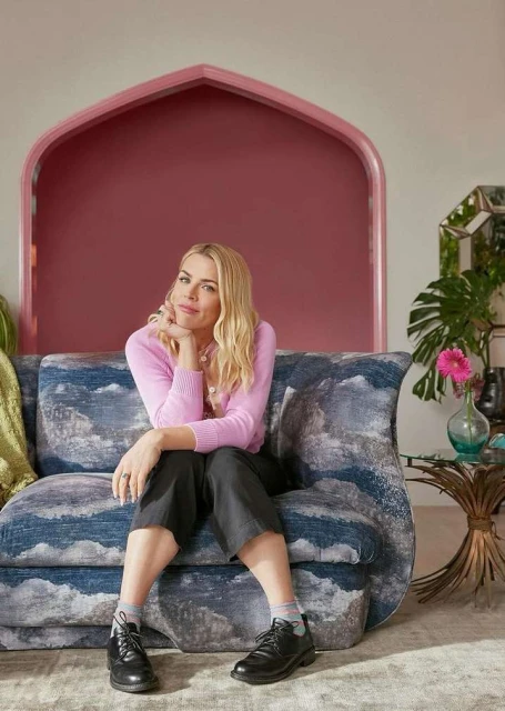 Adult woman with blonde hair sits on a couch in a relaxed pose in an interior setting with plants, table, and colorful decor.