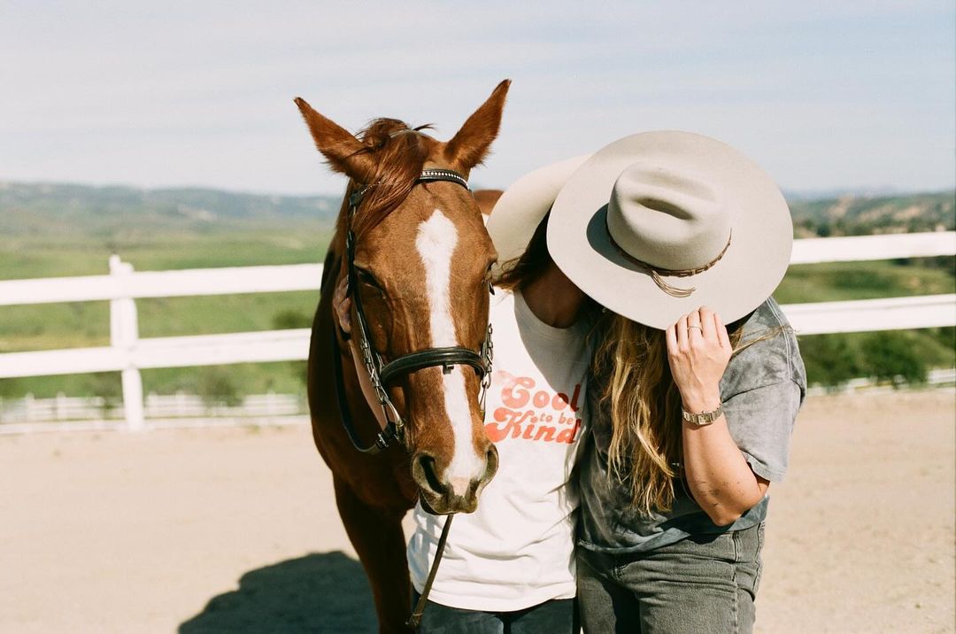 Woman embraces a brown horse in an outdoor paddock with a distant green valley, wearing a cowboy hat and a t-shirt that says, 'Good to be Kind'.