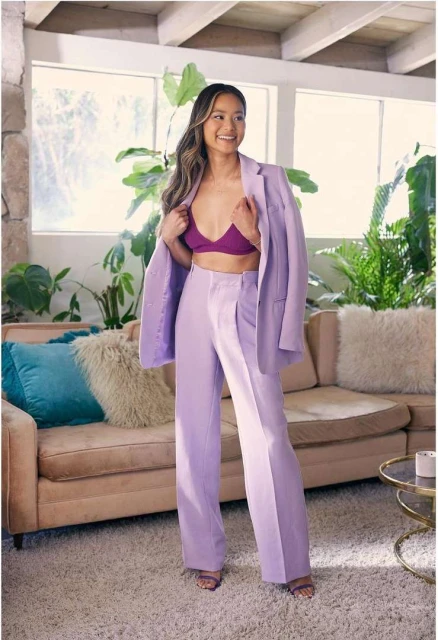 Stylish woman poses in lilac blazer and trousers, smiling in bright indoor setting with lush plants and neutral furnishings.