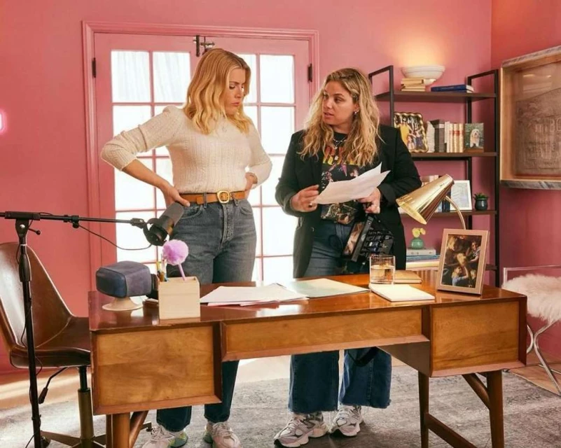 Two women stand at a desk in a pink room, one holding papers, the other with hands on hips, with a microphone and various objects visible.