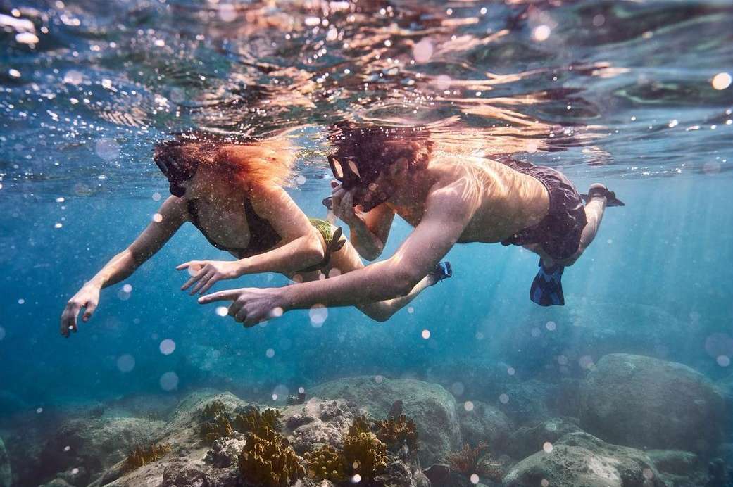 Underwater shot of two snorkelers in the ocean wearing masks and fins surrounded by rocks and clear blue water with sun rays.
