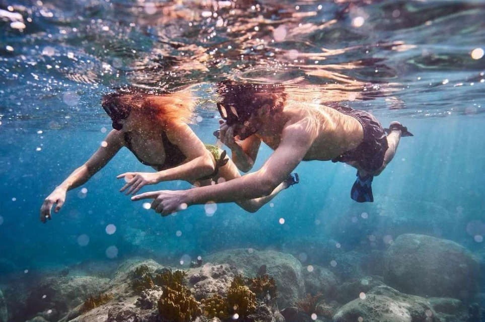 Underwater shot of two snorkelers in the ocean wearing masks and fins surrounded by rocks and clear blue water with sun rays.