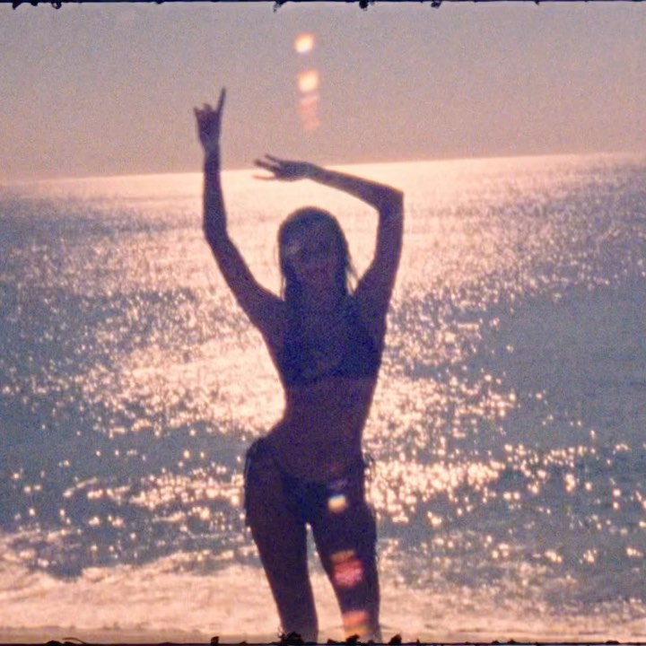 A silhouetted figure of an adult female stands on a sandy beach at sunset, with the sun reflecting on the rippling water in the background.
