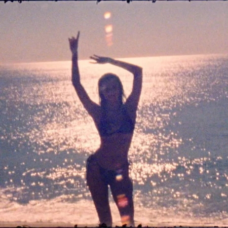 A silhouetted figure of an adult female stands on a sandy beach at sunset, with the sun reflecting on the rippling water in the background.
