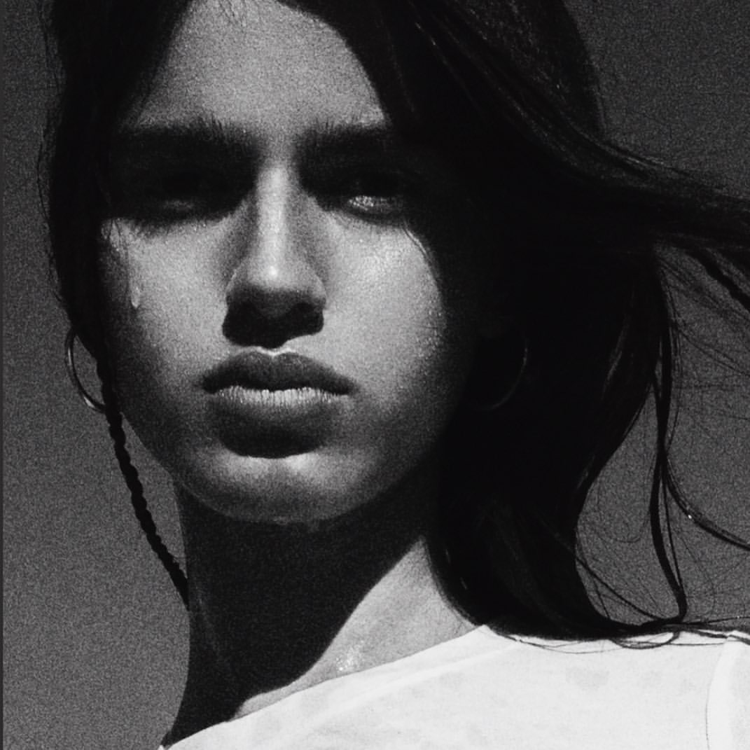 Close-up portrait of a young adult woman with dark hair, wearing a white t-shirt, with a tear on her cheek in a black and white photograph.