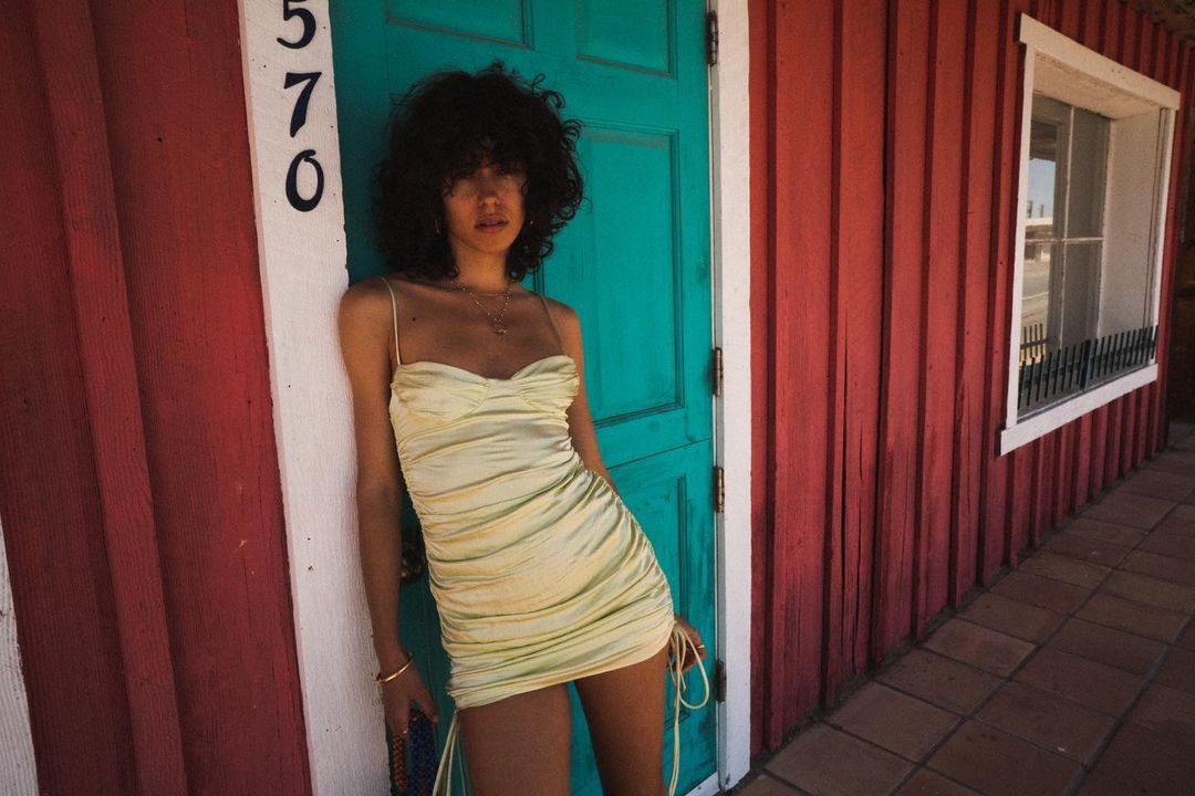 A young woman with curly hair poses in a yellow dress outdoors. The shot features a vibrant turquoise door and a red wall.