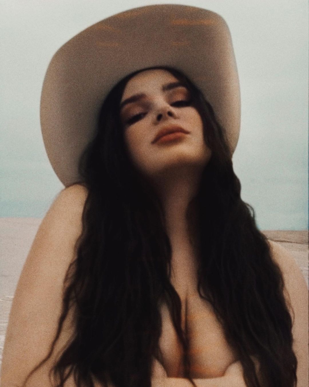Portrait of woman with long dark hair wearing a cream-colored cowboy hat against a sky and beach background.
