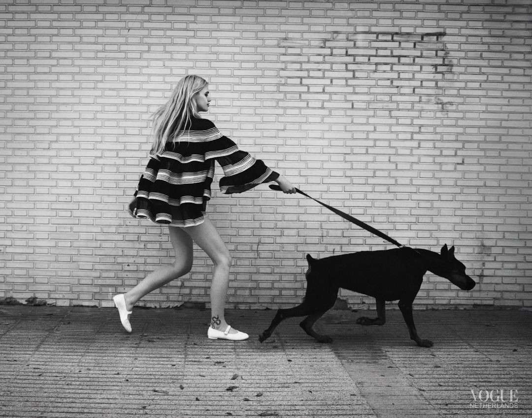 A person walks a black dog on a leash on a sidewalk in front of a brick wall in a black and white fashion shot, capturing movement and style.