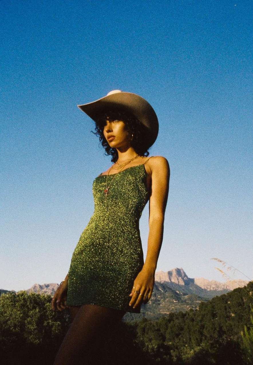 A woman with curly hair in a green dress and cowboy hat poses outdoors with a mountain backdrop under a bright blue sky.