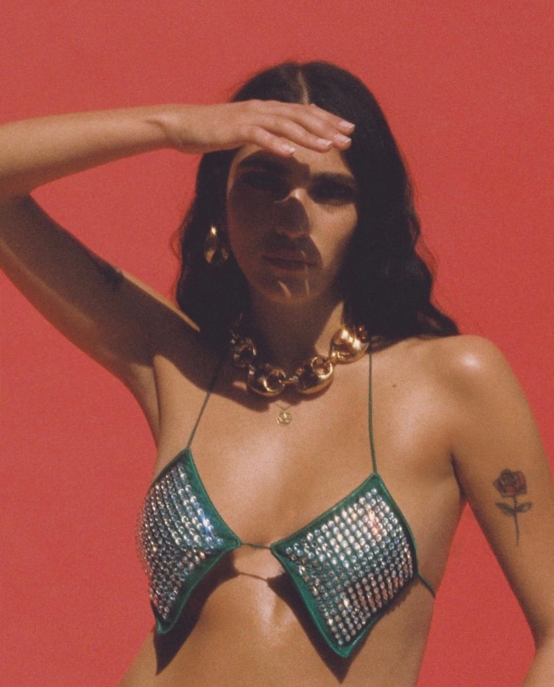 A woman poses against a red background, wearing a jeweled green bra top, a gold necklace, and gold hoop earrings, shading her eyes.