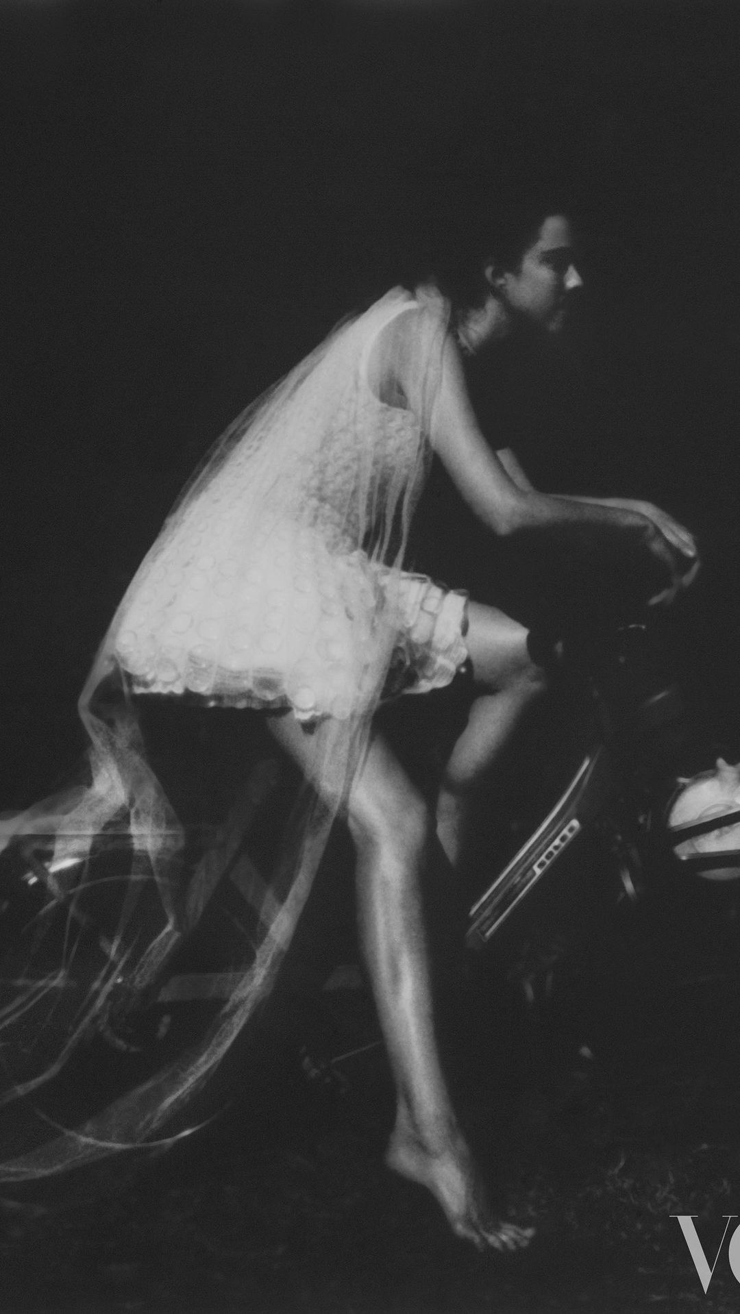 Black-and-white fashion photograph of an adult female riding a motorcycle with motion blur, lace dress, and Vogue logo.