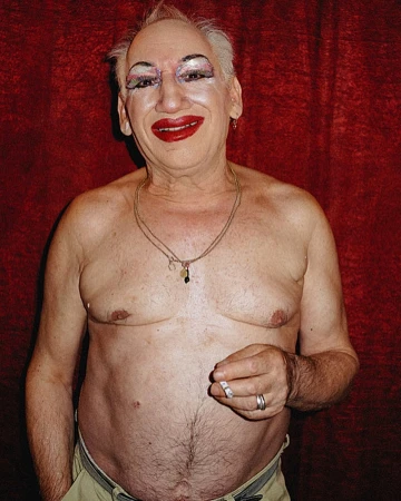 Adult male with heavy makeup poses indoors with a lit cigarette and gold jewelry against a red velvet-textured backdrop.