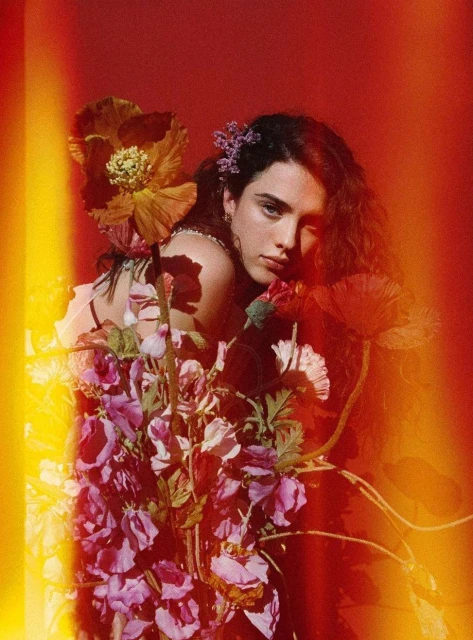 Portrait of a woman surrounded by colorful flowers in a warm-toned, photo studio setting with a red backdrop and bright highlights.