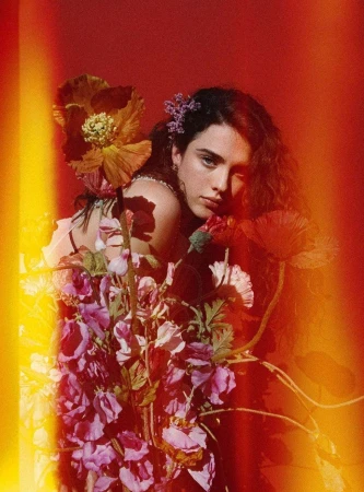 Portrait of a woman surrounded by colorful flowers in a warm-toned, photo studio setting with a red backdrop and bright highlights.
