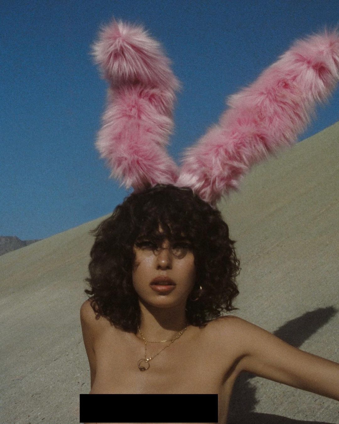 Woman with bunny ears poses against a desert landscape under a clear sky, showcasing fashion and beauty elements.