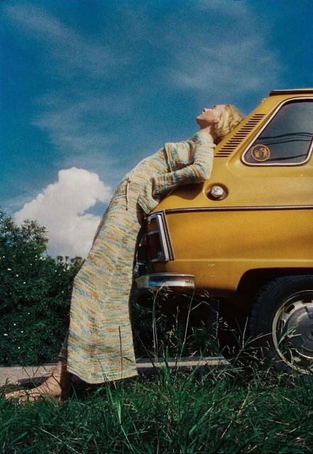 Adult female in a long knit dress leans against the trunk of a yellow car under a bright blue sky in a rural setting.