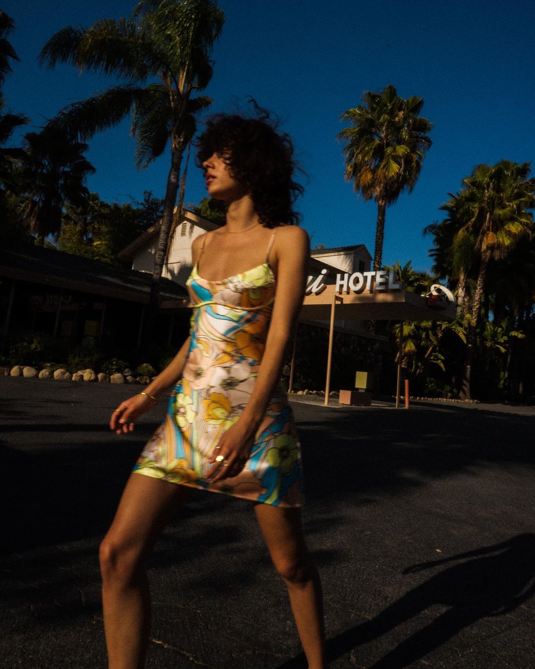 Woman in floral dress stands near hotel under blue sky with palm trees, bathed in warm sunlight, creating a vintage-inspired fashion scene.