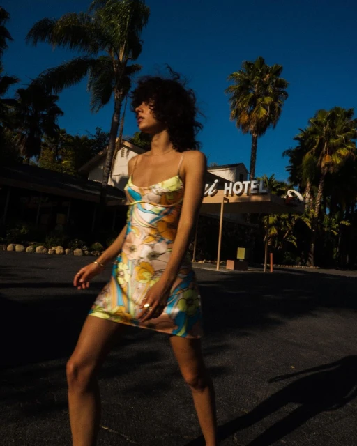 Woman in floral dress stands near hotel under blue sky with palm trees, bathed in warm sunlight, creating a vintage-inspired fashion scene.