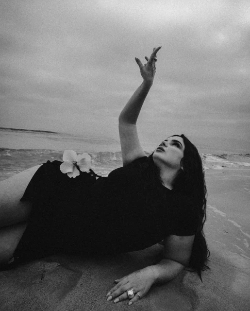 Woman lying on sandy beach near ocean in black dress and flower accessory raises arm in black and white photograph on cloudy day.