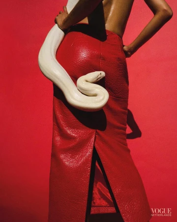 Woman in red skirt poses with a large white snake against a vibrant red background in a fashion editorial shot. Vogue logo visible.