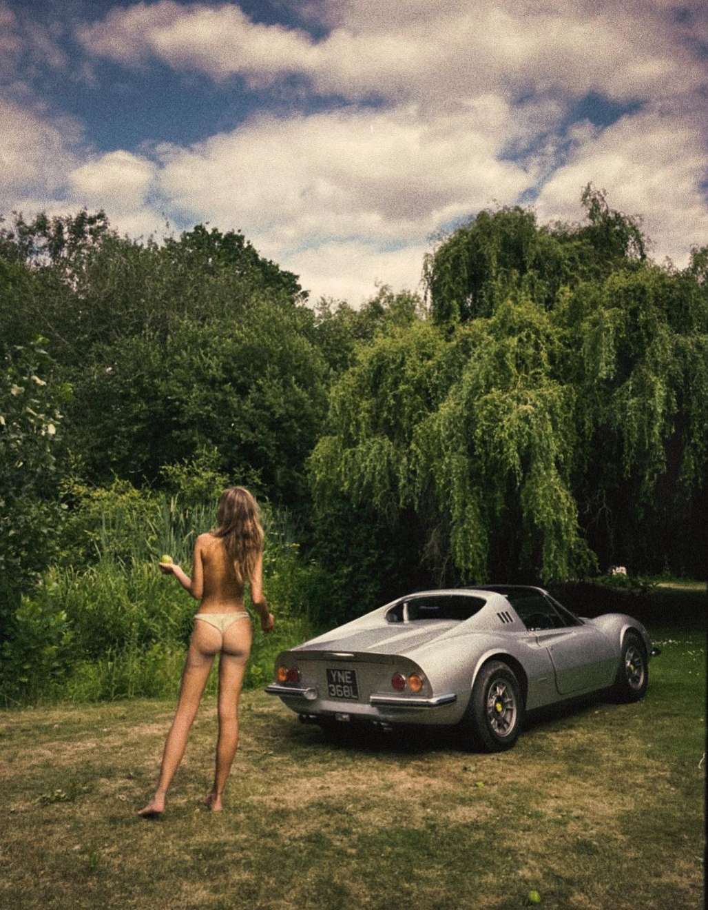 A woman in swimwear stands near a silver vintage sports car in a grassy outdoor setting with trees and a cloudy sky above.