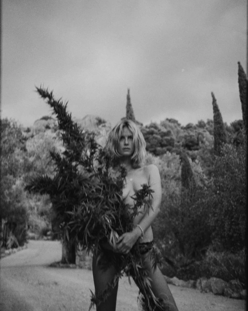 Monochrome portrait of adult female holding a large leafy plant outside, medium shot with trees and cloudy sky in the background.