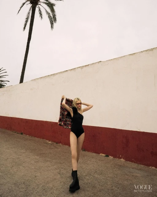 A blonde fashion model in a black one-piece and black boots stands on a street, draped with a patterned jacket, next to a palm tree.