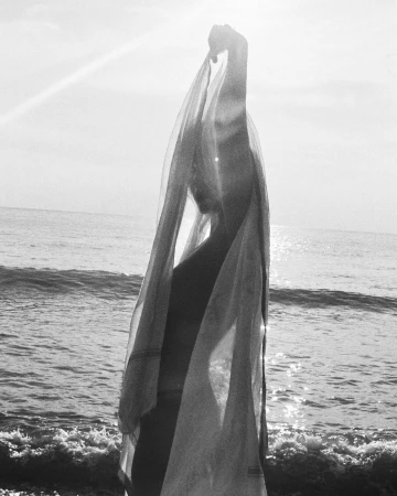 Black and white portrait of a woman holding a sheer fabric overhead on a sandy beach with ocean waves in the background.