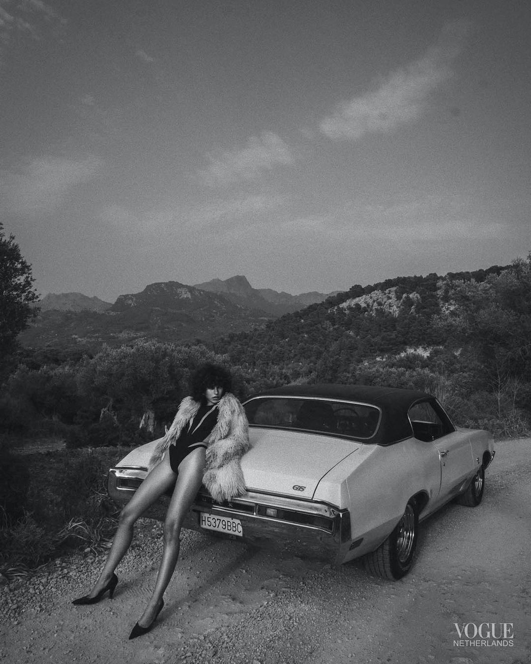 Black-and-white shot of a woman in a bodysuit and fur coat leaning against a vintage car in a mountainous rural setting.